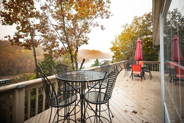 A wooden deck with a round table and chairs, overlooking a lake with autumn foliage.