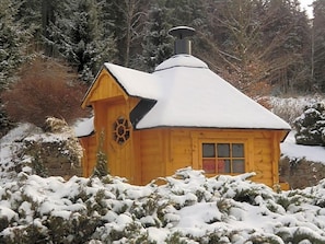 Une cabane en bois dont le toit est recouvert de neige, entourée d’arbustes et d’arbres chargés de neige.