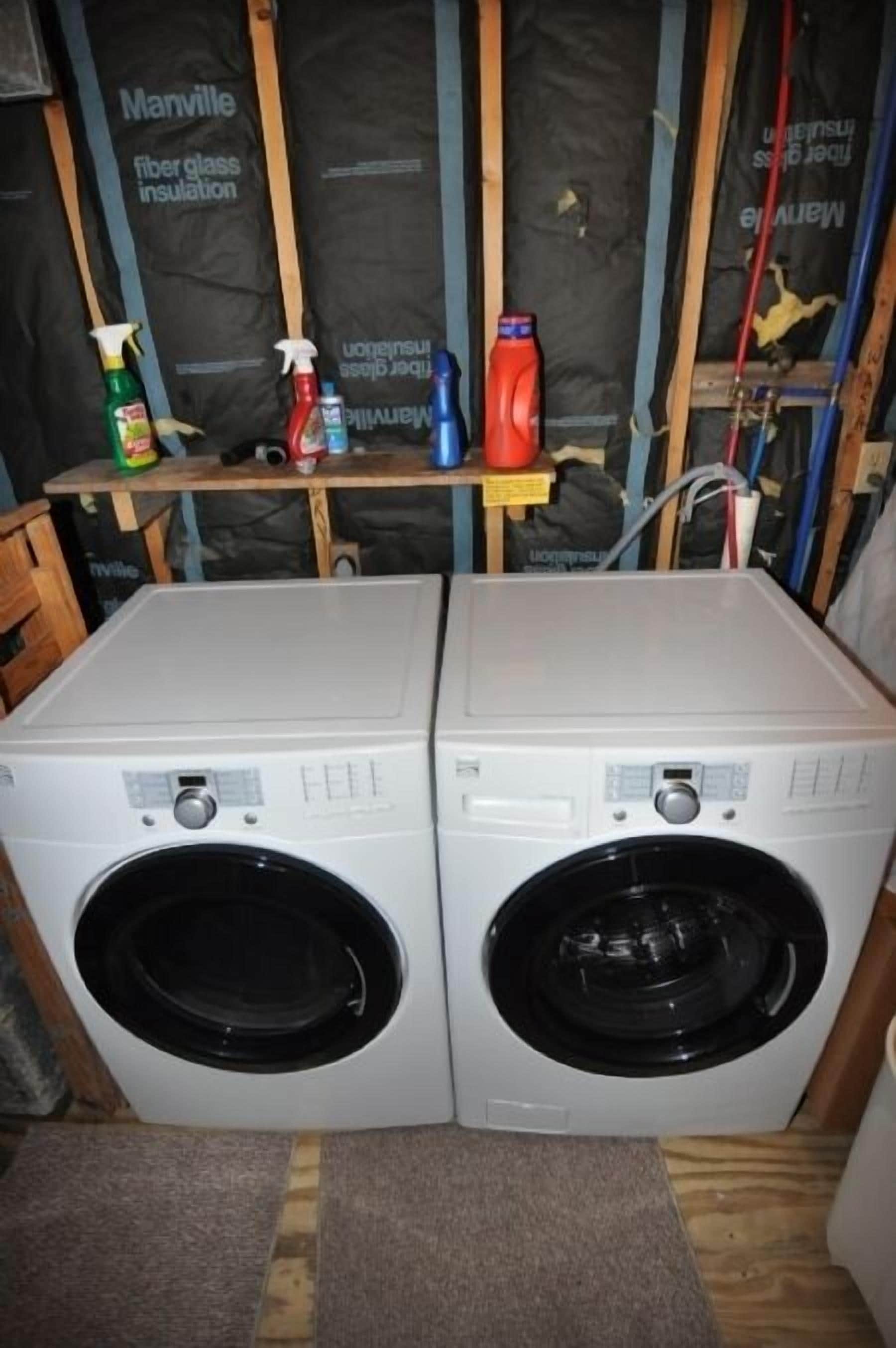 Two front-loading washing machines in a laundry area with insulation panels and cleaning supplies on a shelf.