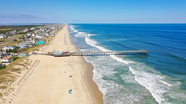 Una playa con un muelle que se adentra en el océano, rodeada de edificios residenciales.