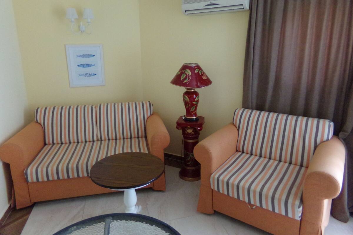 A room with two striped sofas, a small round table, a red lamp, an air conditioning unit, and a framed picture on the wall.