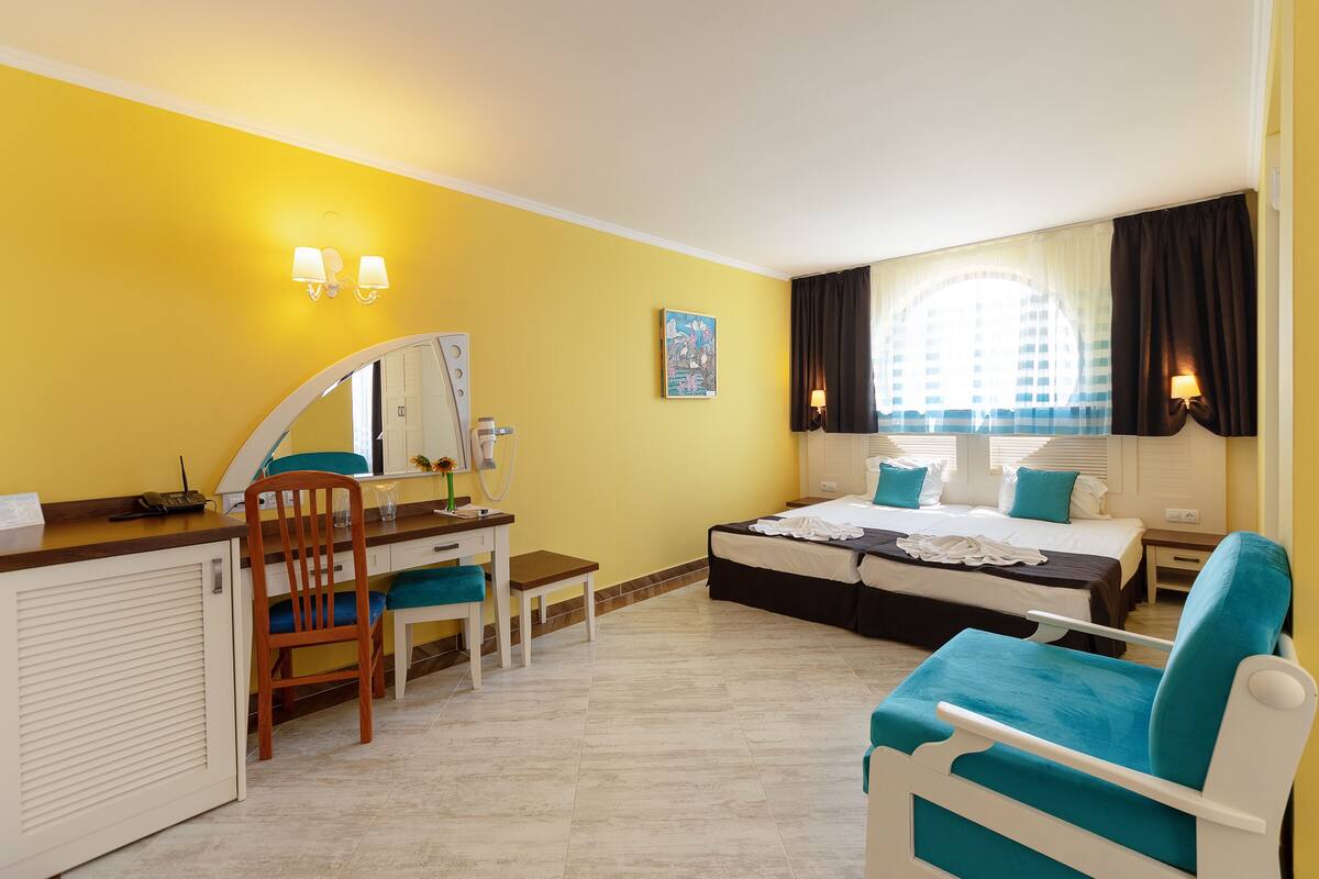 A hotel room with a bed, a desk with a chair, a mirror, a window with curtains, and a painting on the wall.