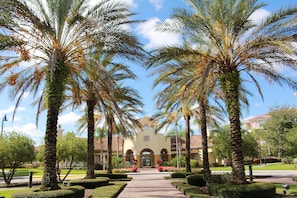 Stroll through the beautifully manicured courtyard, surrounded by palm trees and hedges, towards the classic building with a central entrance and arched windows.
