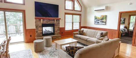 A spacious living room with a stone fireplace, a ceiling fan, and large windows.