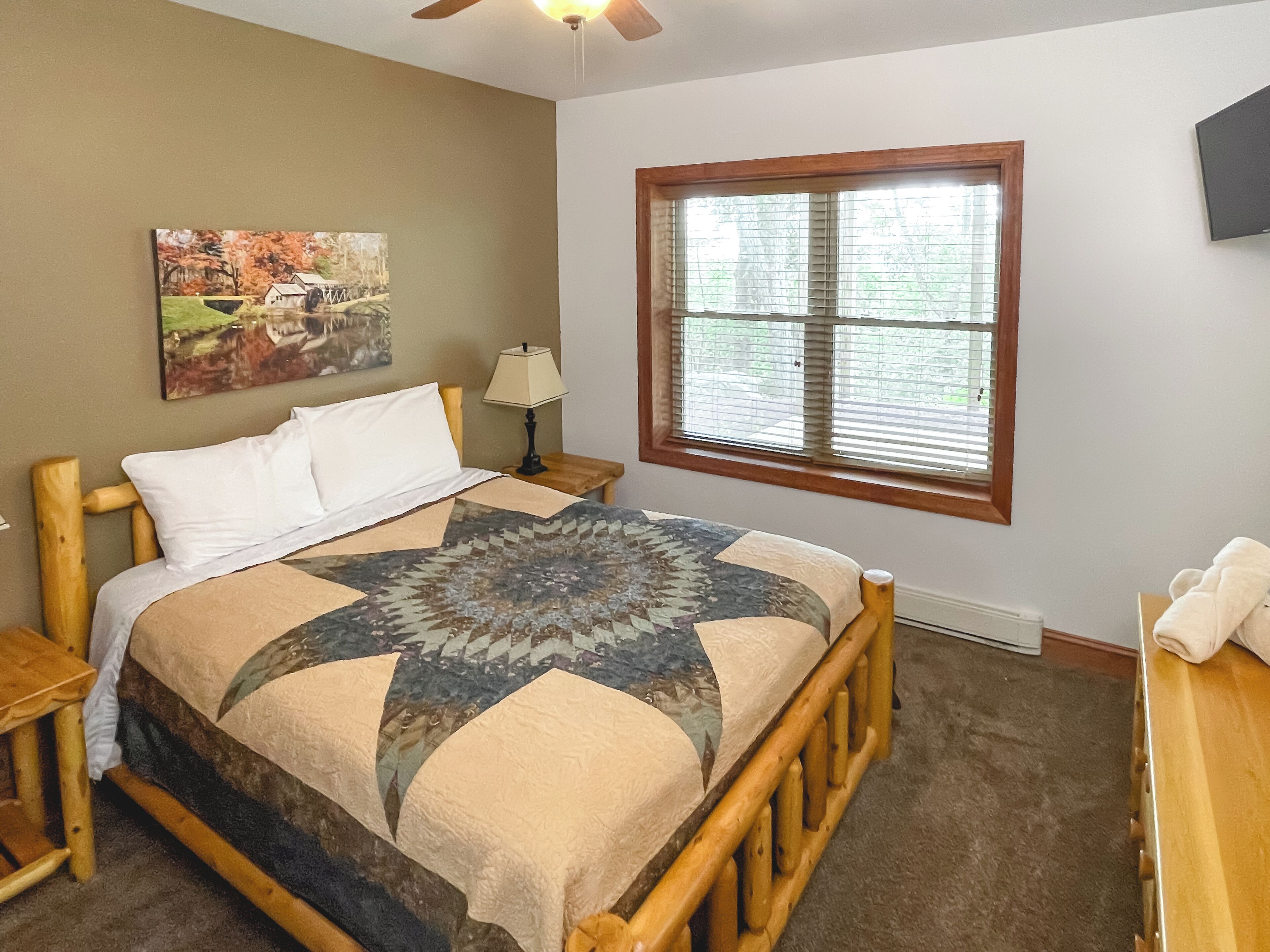 A bedroom with a wooden bed, a window with blinds, a painting on the wall, and a lamp on a nightstand.
