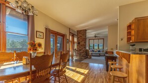 A spacious kitchen with wooden flooring, a dining area with wooden chairs, a brick fireplace, and a large window with curtains.