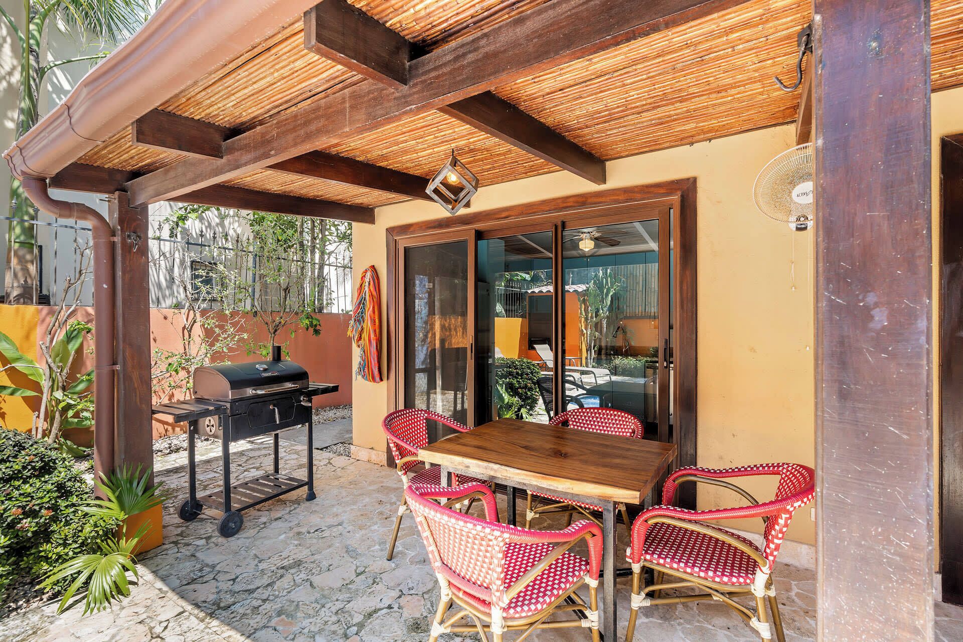 An outdoor patio with a wooden table and wicker chairs, a grill, and a glass door leading to a covered area.