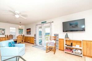 A living room with a television, a dining area with chairs, a rocking chair, and a bookshelf with books and a model ship. - What Time Is It? - Quaint 3 BR Home - Check in any day! (Surf City)
