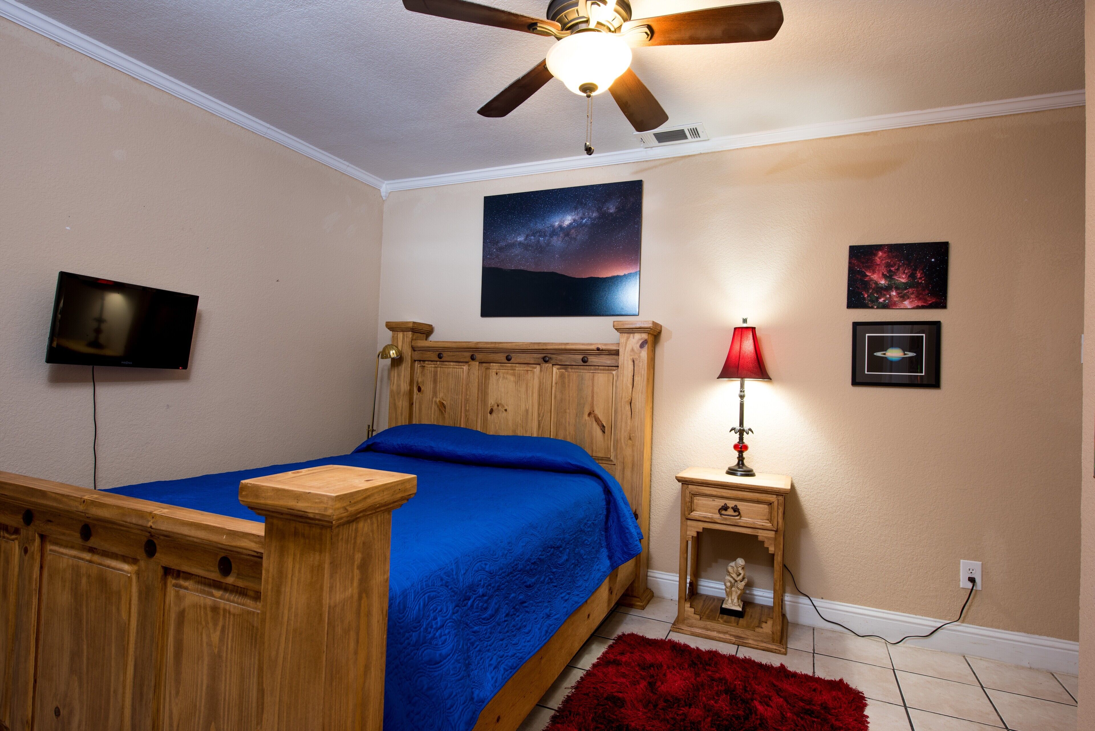 Une chambre à coucher avec un lit en bois, un ventilateur de plafond, un téléviseur fixé au mur et une table de chevet avec une lampe.
