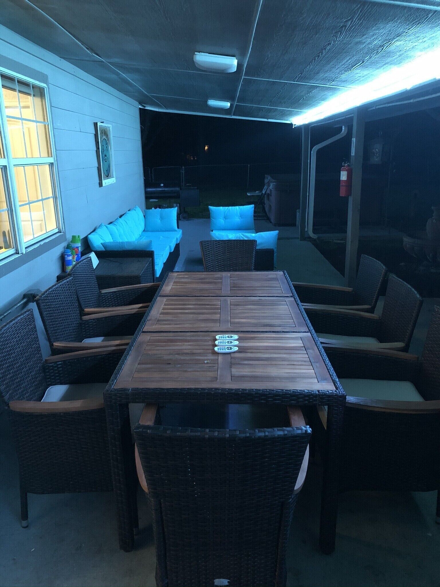 A covered outdoor seating area with a wooden table and wicker chairs, a sofa with blue cushions, and a fire extinguisher mounted on the wall.