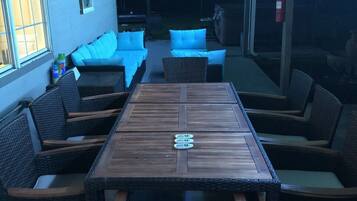 A covered outdoor seating area with a wooden table and wicker chairs, a sofa with blue cushions, and a fire extinguisher mounted on the wall.