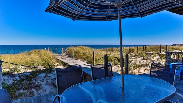 A beachside patio with a glass table and chairs, a striped umbrella, and a view of the ocean. - Waterfront cottage with private beach in Sandwich! 179B North Shore Blvd (Sandwich)
