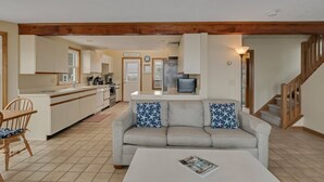 A modern kitchen with white cabinets, a built-in oven, and a refrigerator. There is a dining area with a wooden table and chairs. A sofa with star-patterned cushions is in the living room. A staircase leads to another level. - Waterfront cottage with private beach in Sandwich! 179B North Shore Blvd (Sandwich)