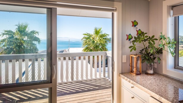 A balcony with a wooden floor, a white railing, and a view of the ocean and palm trees. - THE WAIHEKE PAD | Be My Guest (Oneroa)