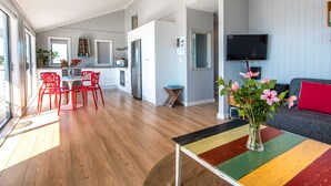 A modern kitchen with white cabinets, a dining area with red chairs, and a living room with a colorful table and a TV. - THE WAIHEKE PAD | Be My Guest (Oneroa)