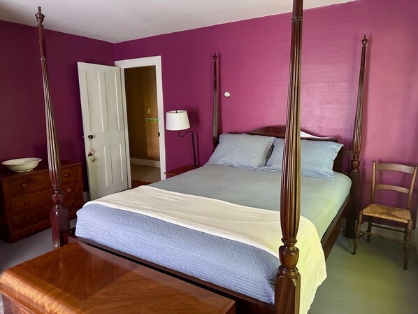 Una cama con dosel, cabecera y piecera de madera, una mesita de noche con un cuenco, y una silla.