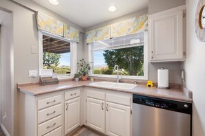 A kitchen with white cabinets, a stainless steel dishwasher, a sink, and a window with a view of greenery and a distant landscape. - Peaceful Sports Village Retreat | Beautiful Mountain Views, Pools & Patio (St George)