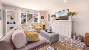 A living room with a sofa, armchair, ottoman, and a flat-screen TV mounted on a white cabinet. - Peaceful Sports Village Retreat | Beautiful Mountain Views, Pools & Patio (St George)