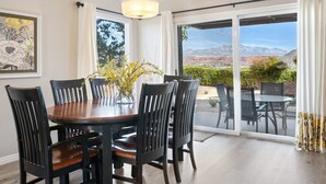 A dining area with a wooden table and chairs, a vase with yellow flowers, and a view of mountains through large sliding glass doors. - Peaceful Sports Village Retreat | Beautiful Mountain Views, Pools & Patio (St George)