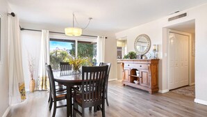 A dining area with a wooden table and chairs, a wooden cabinet with a mirror, and a view of the outdoors through large windows. - Peaceful Sports Village Retreat | Beautiful Mountain Views, Pools & Patio (St George)