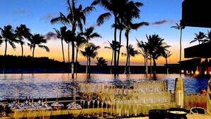 Un bar frente a la playa con vistas a palmeras y al océano al atardecer.