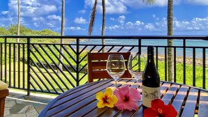 Una mesa con una botella de vino, dos copas y flores en un balcón con vista al océano y palmeras.