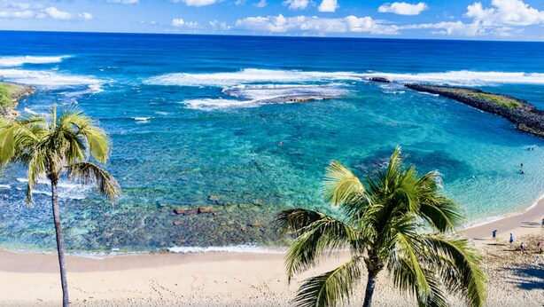 Una playa con agua turquesa cristalina, palmeras y afloramientos rocosos.