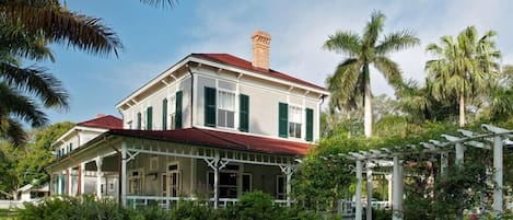 A two-story house with a red roof and green shutters, surrounded by lush greenery and palm trees.