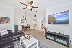 A modern living room with a flat-screen TV, a ceiling fan, a dining area, and a kitchen in the background. - Summerlin Woods - Second floor condo (Fort Myers)