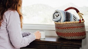 Eine Person sitzt am Fenster mit einem gewebten Korb, der eine aufgerollte Decke und eine Tasse enthält.