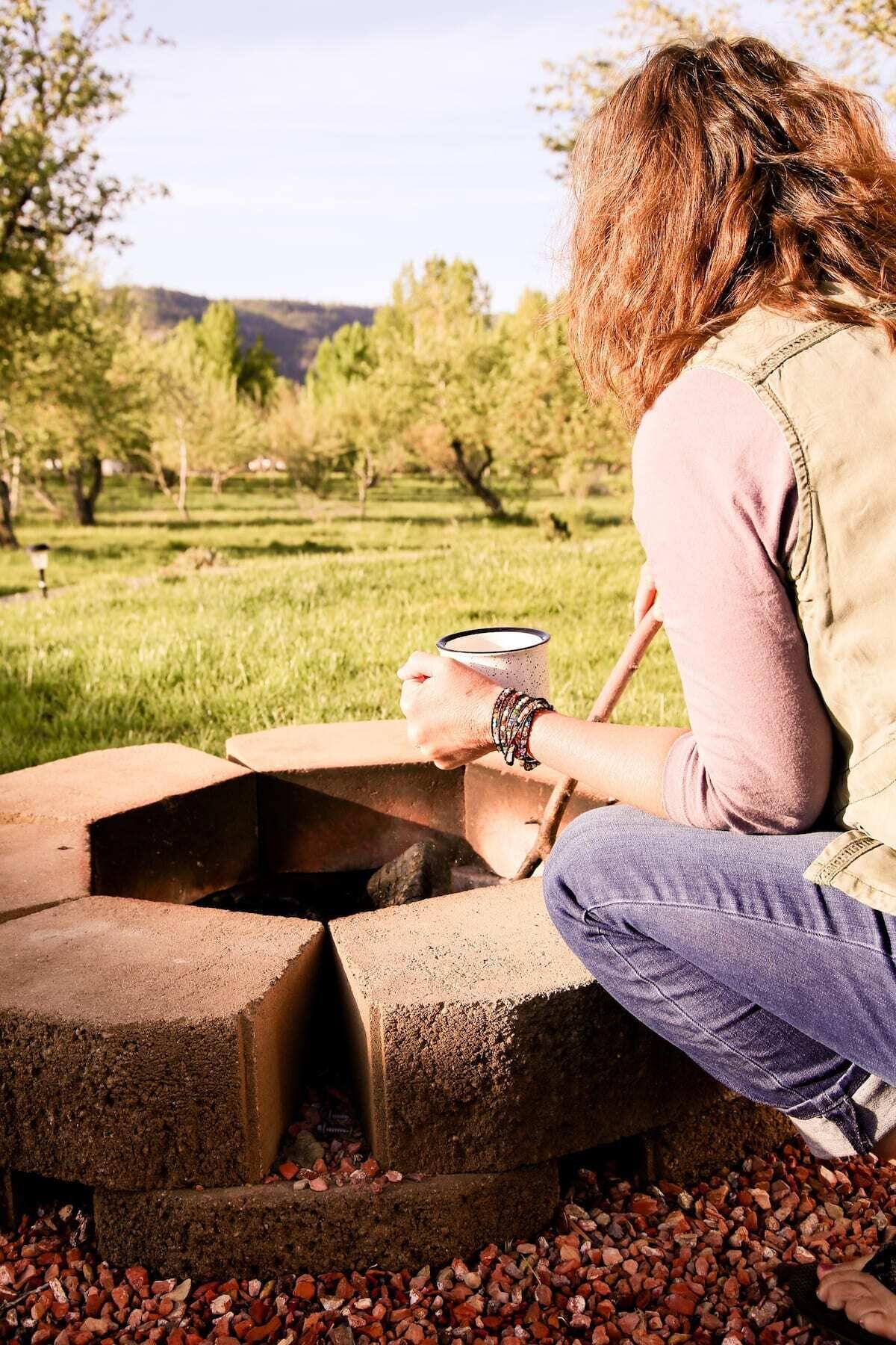 Una persona sentada junto a una fogata improvisada al aire libre, sosteniendo una taza.