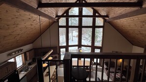 A high-ceilinged room with wooden beams, a large window with a view of a snowy landscape, a dining table with chairs, and a staircase leading to an upper level. - Ski-in Ski-out Killington Chalet on Great Eastern Trail, Huge Loft + 4 Bedrooms (Killington)