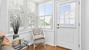 A room with a white door, a wicker chair with a cushion, a small table with a vase and a book, and large windows with blinds.