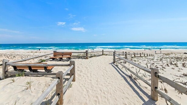 A wooden boardwalk leading to a sandy beach with benches and a wooden fence.