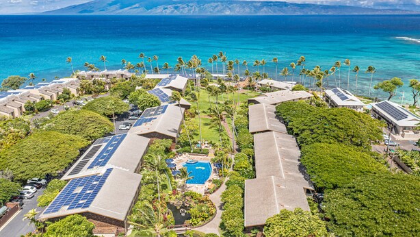 An aerial view of a resort with multiple buildings, solar panels, a swimming pool, and a view of the ocean.