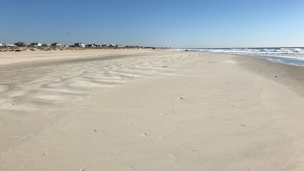A wide sandy beach with footprints leading to the water, a clear sky, and houses in the background. - Prior Engagement: Pet Friendly Beach Cottage at Emerald Isle on The Point (Emerald Isle)