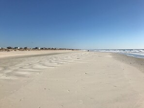 A wide sandy beach with footprints leading to the water, a clear sky, and houses in the background.
