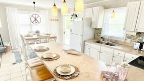 A modern kitchen with white cabinets, a central island with a dining set, pendant lights, and a view of the backyard. - Prior Engagement: Pet Friendly Beach Cottage at Emerald Isle on The Point (Emerald Isle)