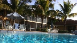 Ein klares Schwimmbecken mit Liegestühlen und Sonnenschirmen, umgeben von Palmen und einem Gebäude mit Balkon.