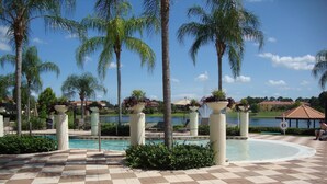 Zona de piscina con baldosas de diseño cuadriculado, palmeras y vistas a un lago y edificios al fondo.