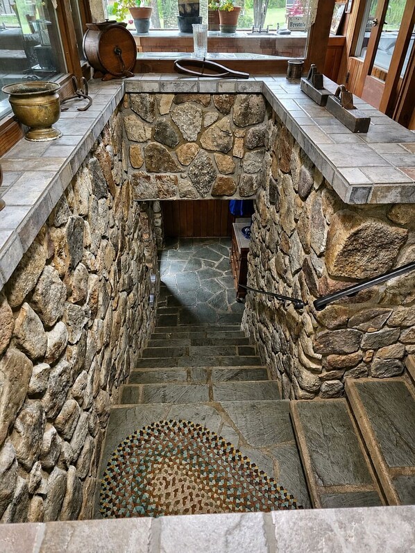 A stone fireplace with a hearth, a stone staircase leading down, and a window with a view of the outdoors. - Brech Mountain House-Hudson Valley (Pine Bush)