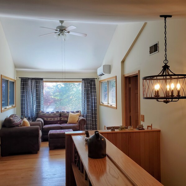 A spacious living room with a ceiling fan, a dining area with a wooden table, and a kitchen area with a hanging light fixture.
