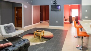 Ein modernes Wohnzimmer mit einer Couch, einem kleinen Tisch und einem an der Wand befestigten Fernseher. Es gibt einen Essbereich mit einer Bar und orangefarbenen Hocker.