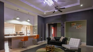 Ein modernes Wohnzimmer mit einer schwarzen Ledersofa, einem weißen Sessel und zwei orangefarbenen Barhocker. In der Mitte steht ein kleiner Holztisch. Der Raum verfügt über einen Deckenventilator und Einbaulicht.