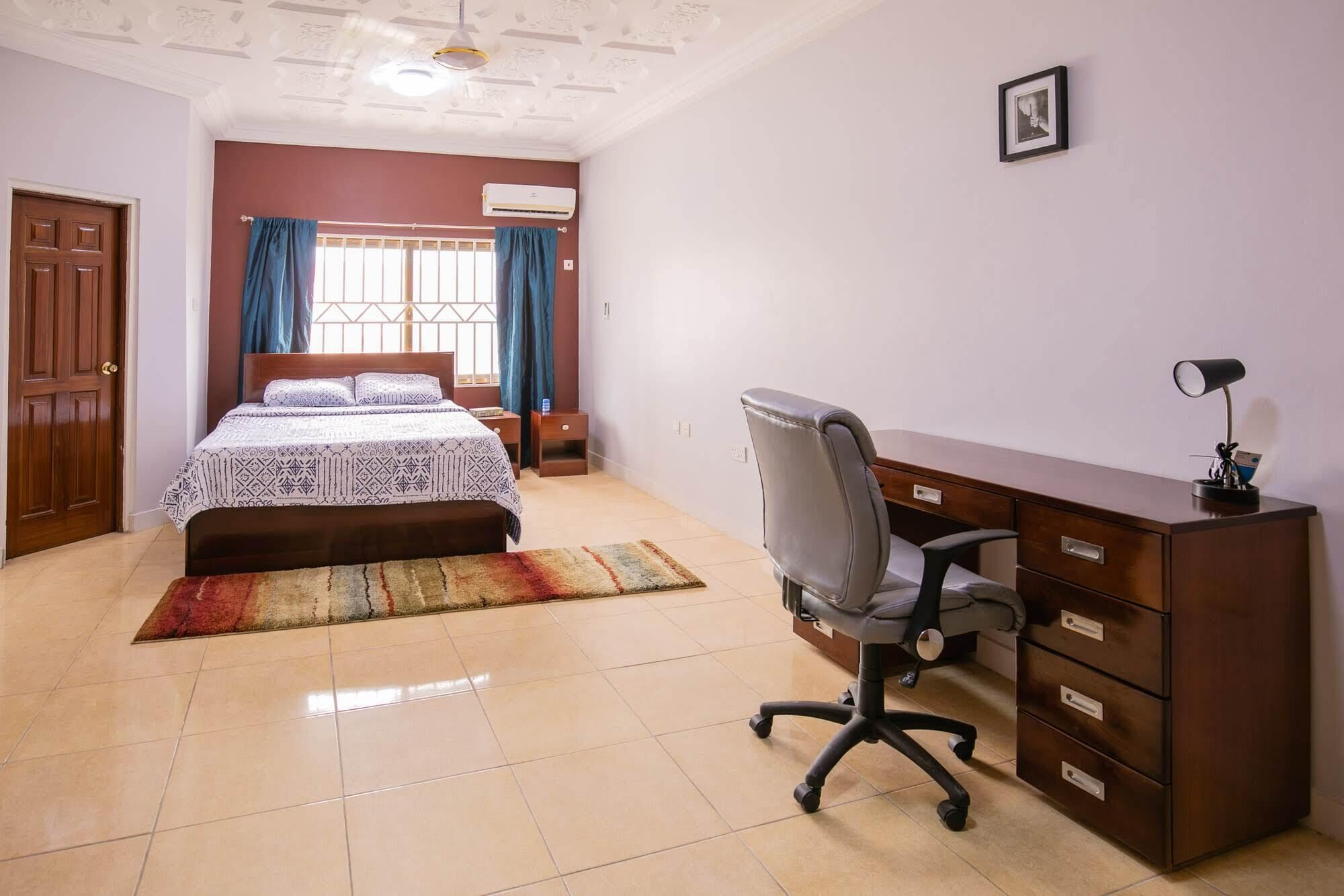 Un dormitorio con cama, escritorio, silla, armario y ventana con cortinas.