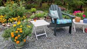 Un jardin aménagé avec un fauteuil Adirondack en bois, une petite table blanche, ainsi que diverses plantes et fleurs en pot.