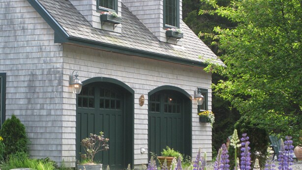 Une maison avec deux portes vertes, un petit porche et un jardin où poussent des fleurs violettes.