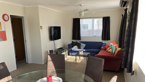 A living room with a glass dining table, a blue sofa, a red sofa, and a television on the wall. - Serenity - our home away from home (Whitianga)