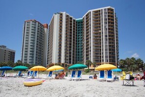 Una playa con edificios altos al fondo, sombrillas y sillones de playa.