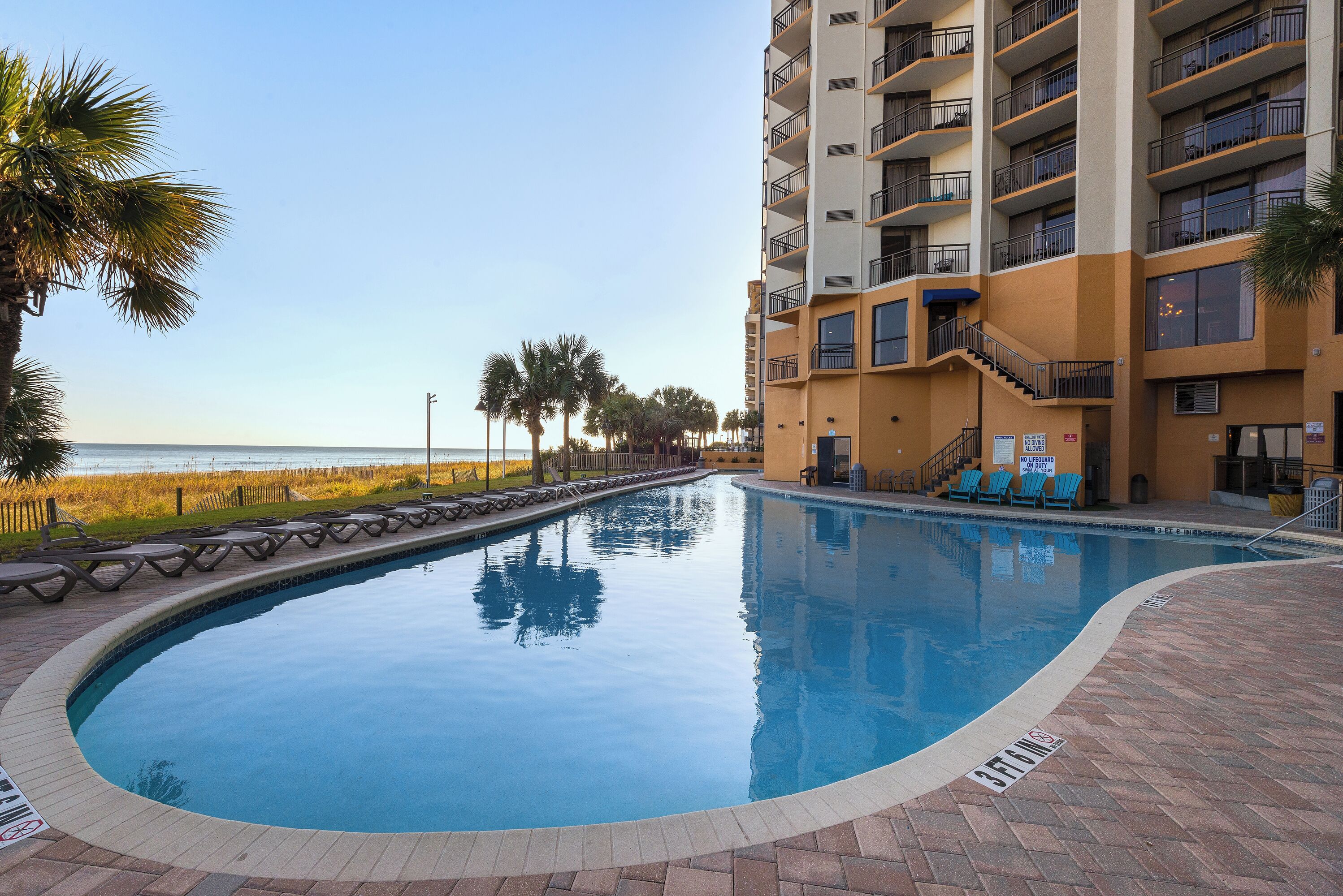 A swimming pool with a curved edge, surrounded by a brick deck. There are lounge chairs and a staircase leading to an upper level. Palm trees and a beach are visible in the background.